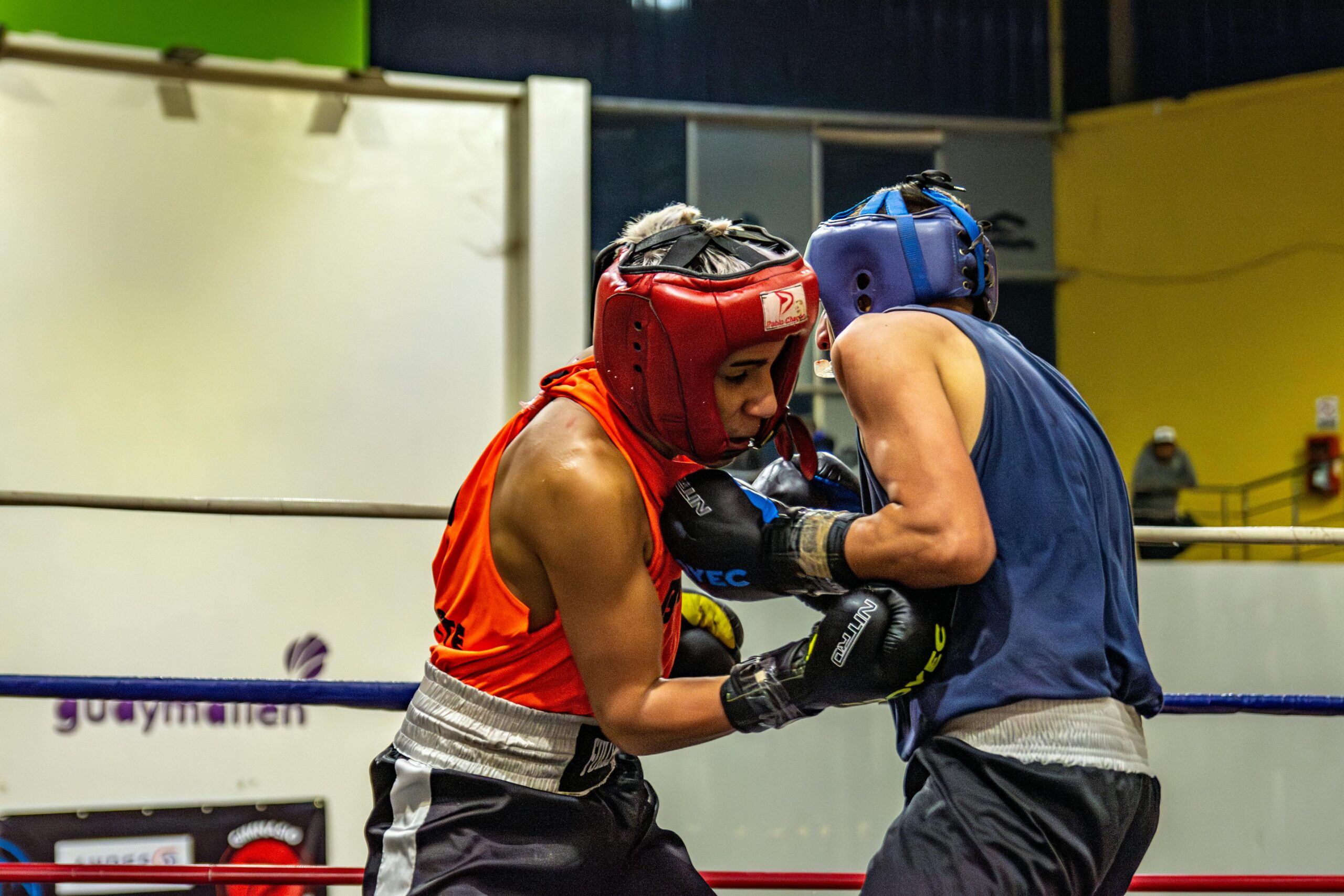 Two athletes in protective gear engage in a spirited boxing match inside a gym.