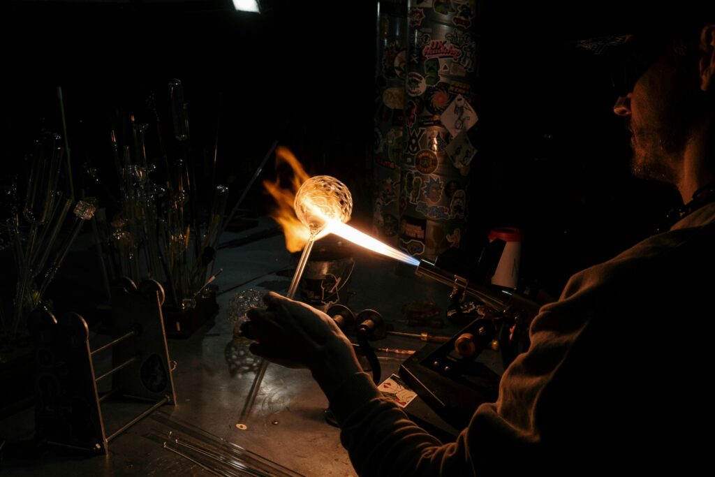 An artisan crafts glass using a blowpipe and flame in a dark workshop setting.