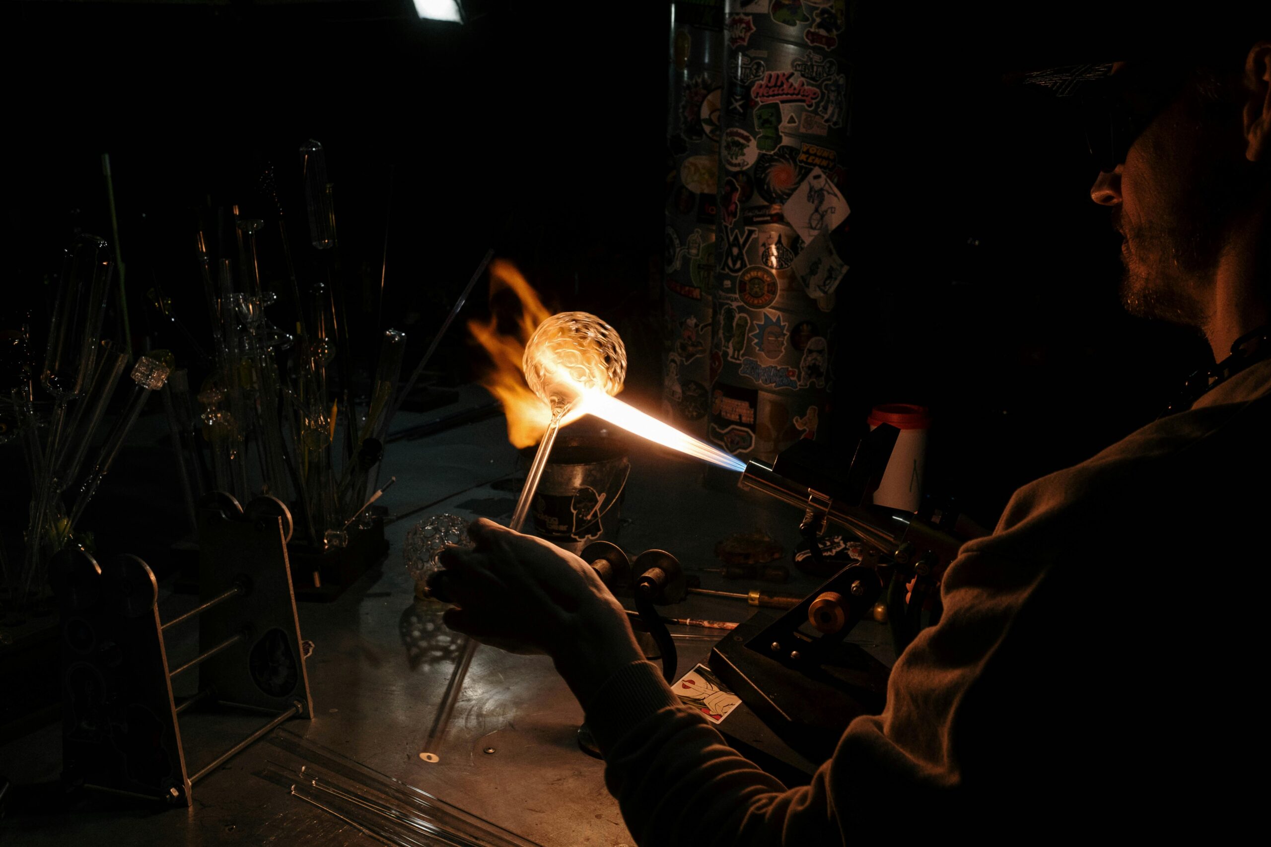 An artisan crafts glass using a blowpipe and flame in a dark workshop setting.